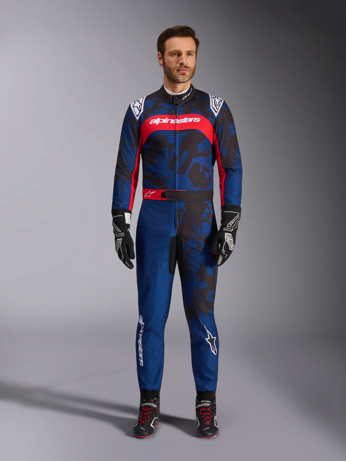 Alpinestars KMX-9 V3 Graphic 5 Suit, combinaison de karting bleu marine et rouge, présentant un motif graphique abstrait de style camouflage, portée par un mannequin avec des gants et chaussures de course noirs, design athlétique avec marquage Alpinestars proéminent sur la poitrine et les jambes.