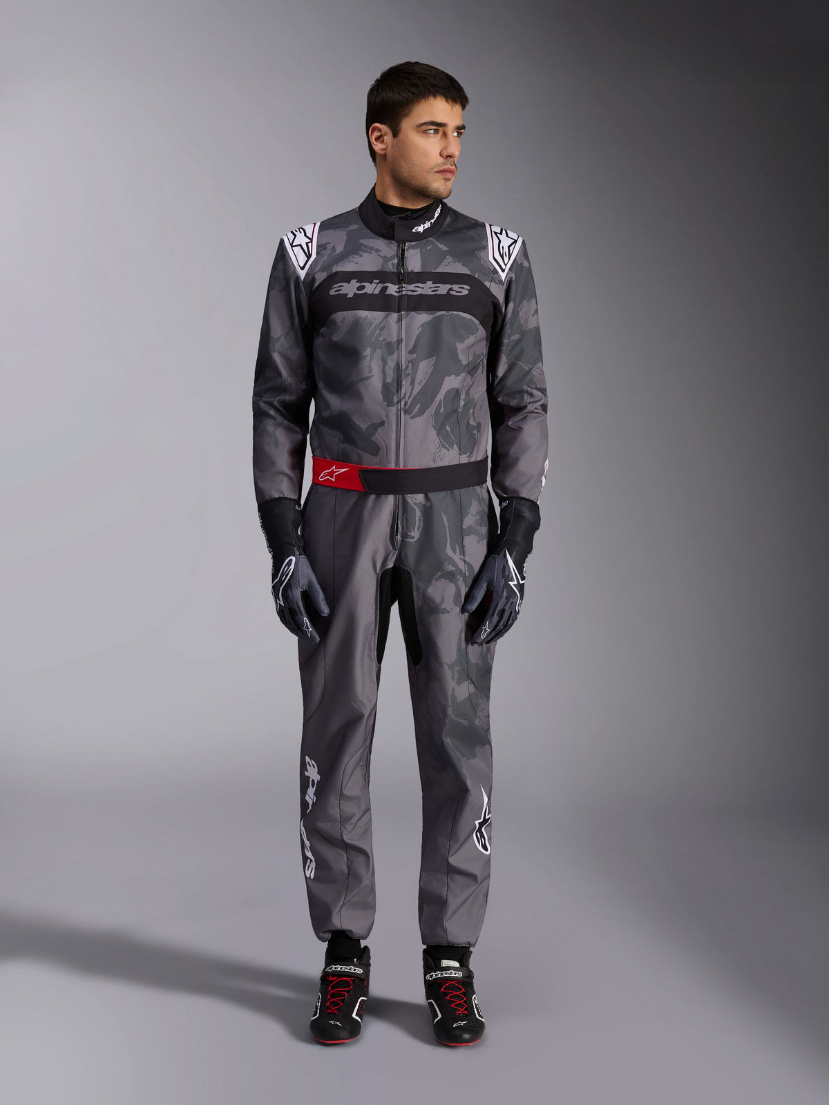 Alpinestars KMX-9 V3 Graphic 5 Anzug, Kartanzug, Dunkelgrau und Schwarz mit Grafiken im Camouflage-Stil, mit schwarzem Brustband mit weißem Logo und rotem Akzent am Taillengürtel, getragen mit passenden Rennhandschuhen und Schuhen, professionelle Motorsportausrüstung