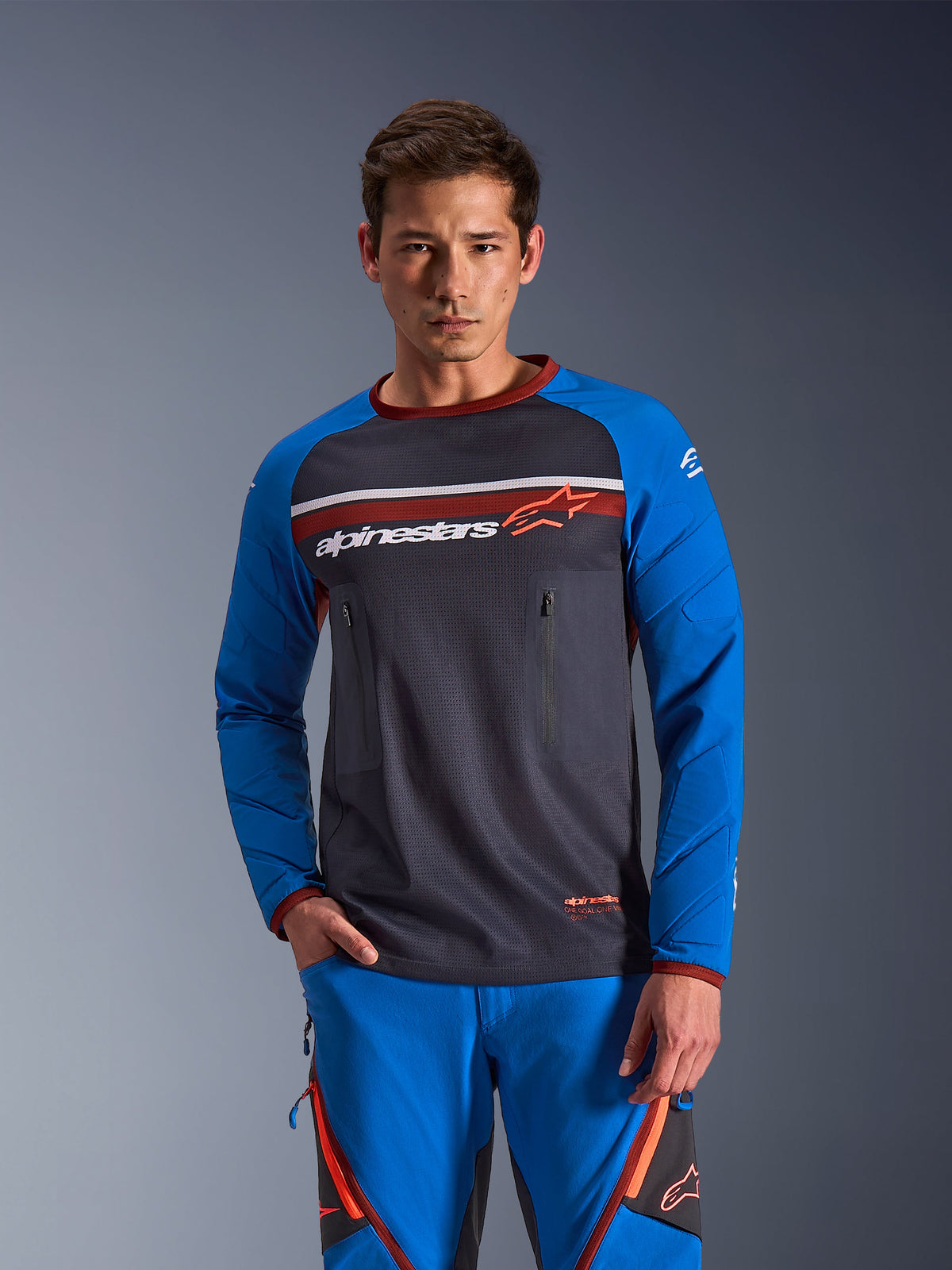Homme portant le maillot Alpinestars Maxdura Dual en gris foncé et bleu royal avec accents orange, torse en maille et poches poitrine zippées, avec pantalon d'aventure bleu assorti, équipement technique pour le dual-sport et le tout-terrain