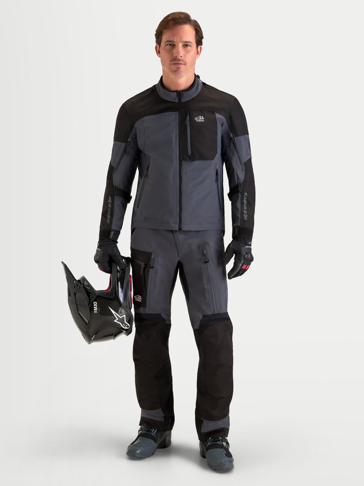 Alpinestars Arenis Gore-Tex Gear Set, adventure motorjas en motorbroek voor heren in zwart en donkergrijs, volledige voorzijde van een model dat staat en een helm vasthoudt