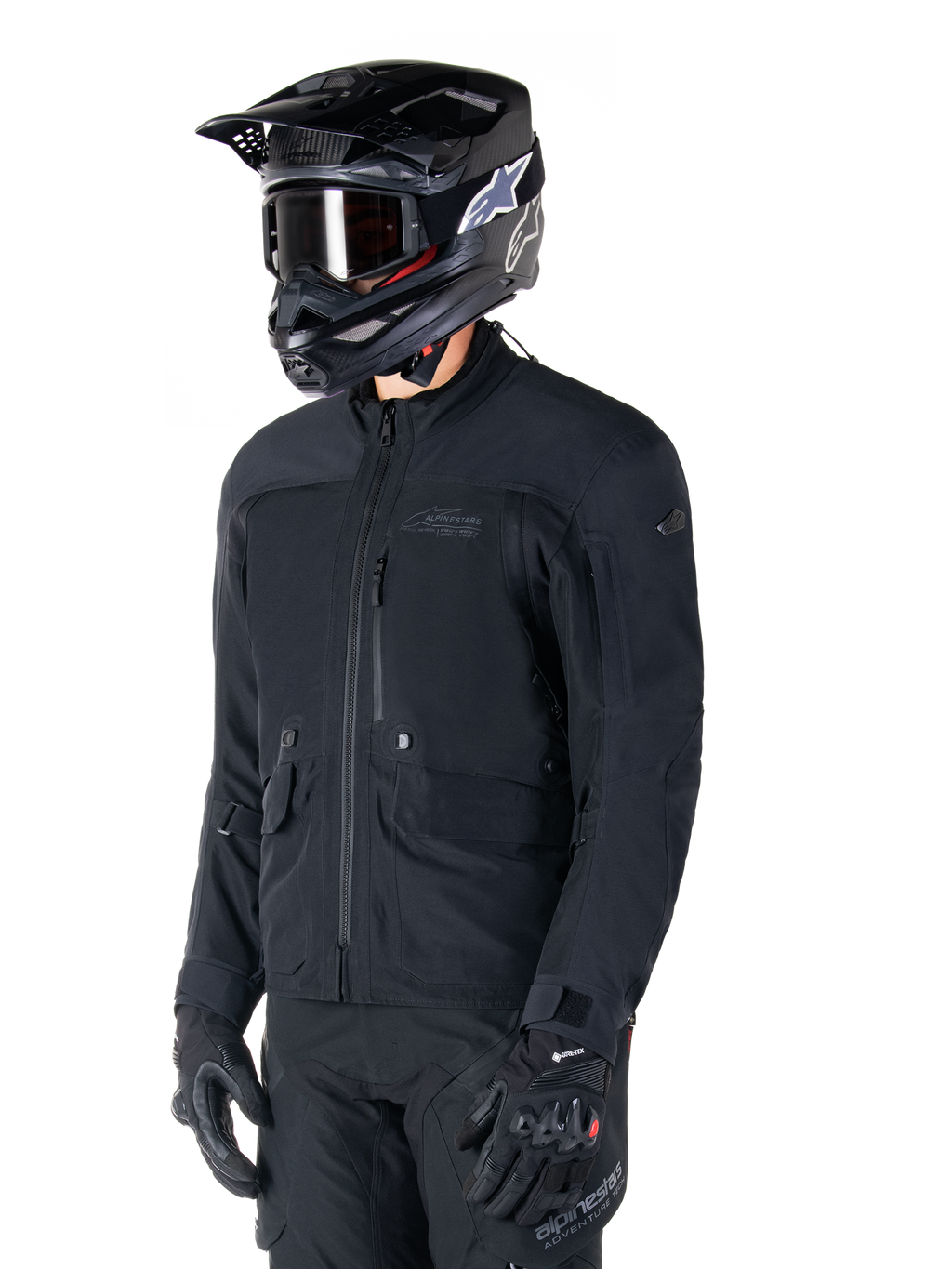 Alpinestars ST-7 Prime GORE-TEX Jacke, schwarze Motorradjacke, getragen von einem Fahrer mit schwarzem Helm und Brille, mit großen Cargo-Taschen auf der Brust und einer laminierten wasserdichten Außenschicht, erstklassige Adventure-Touring- und Pendler-Ausrüstung.