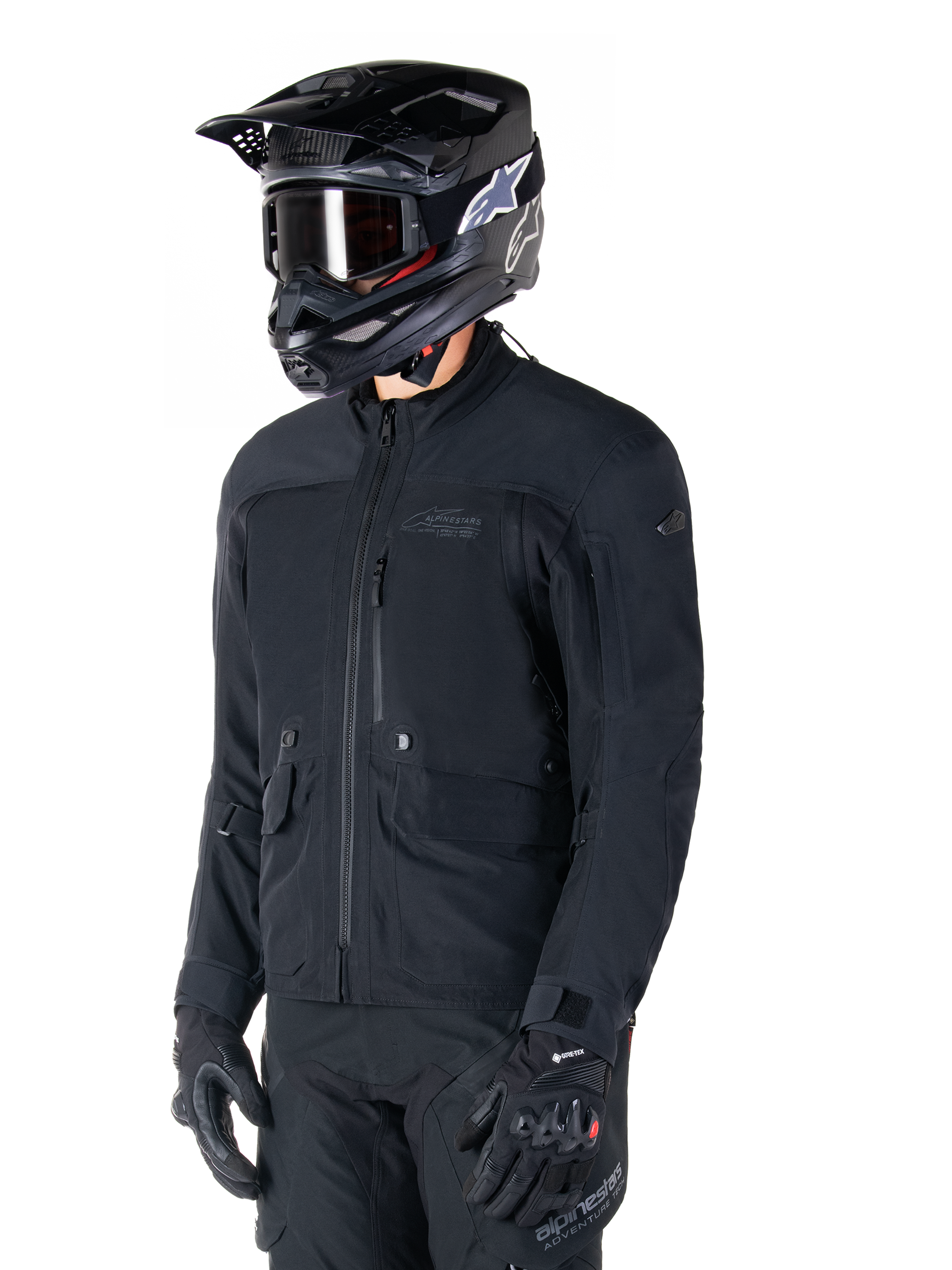 Alpinestars ST-7 Prime GORE-TEX Jacke, schwarze Motorradjacke, getragen von einem Fahrer mit schwarzem Helm und Brille, mit großen Cargo-Taschen auf der Brust und einer laminierten wasserdichten Außenschicht, erstklassige Adventure-Touring- und Pendler-Ausrüstung.
