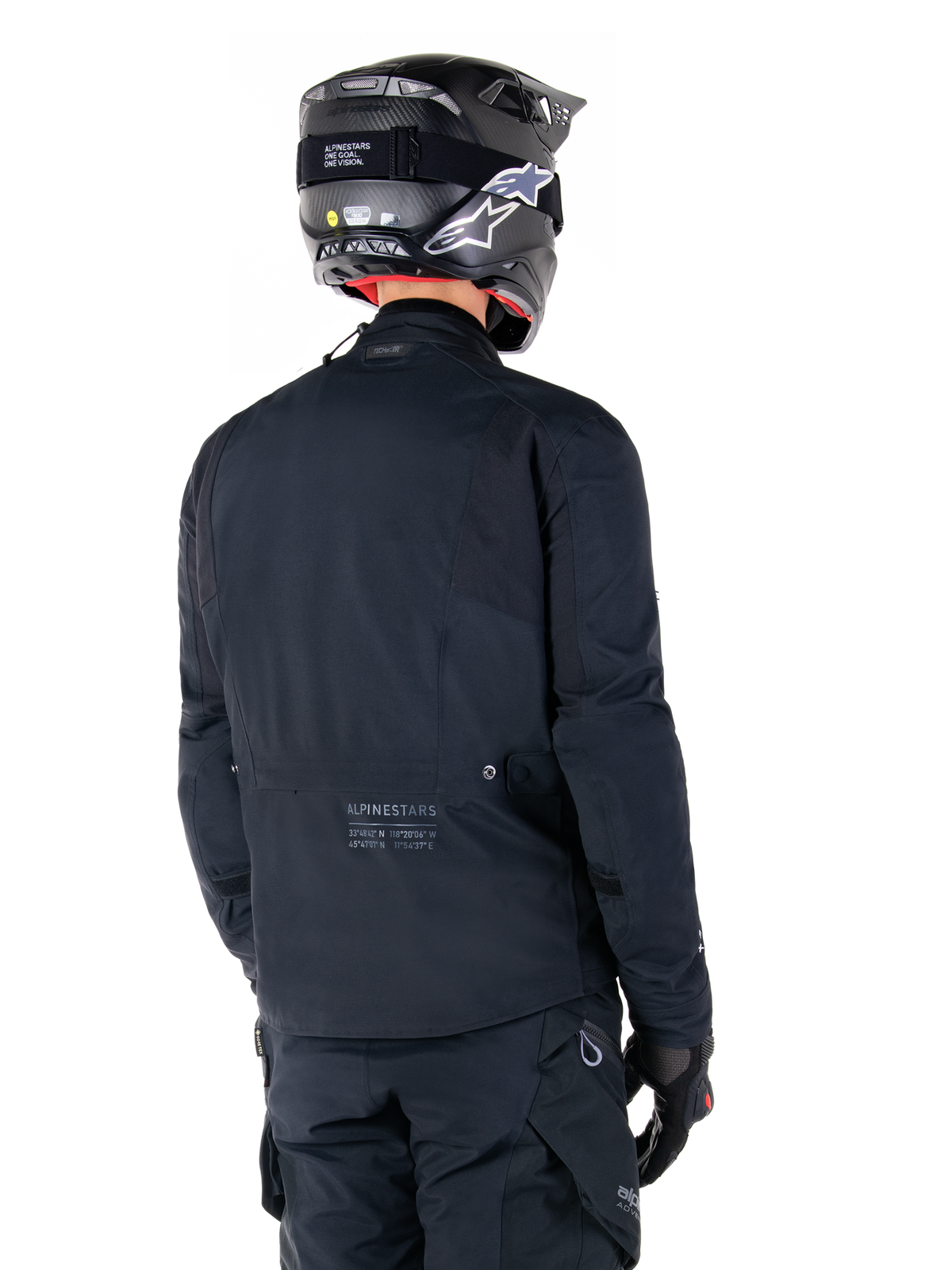 Alpinestars St-7 Prime GORE-TEX Jacke, Schwarz, Rückansicht eines Fahrers, der die Jacke mit passendem Helm und Hose trägt, mit Alpinestars-Branding und geografischen Koordinaten am unteren Rücken.