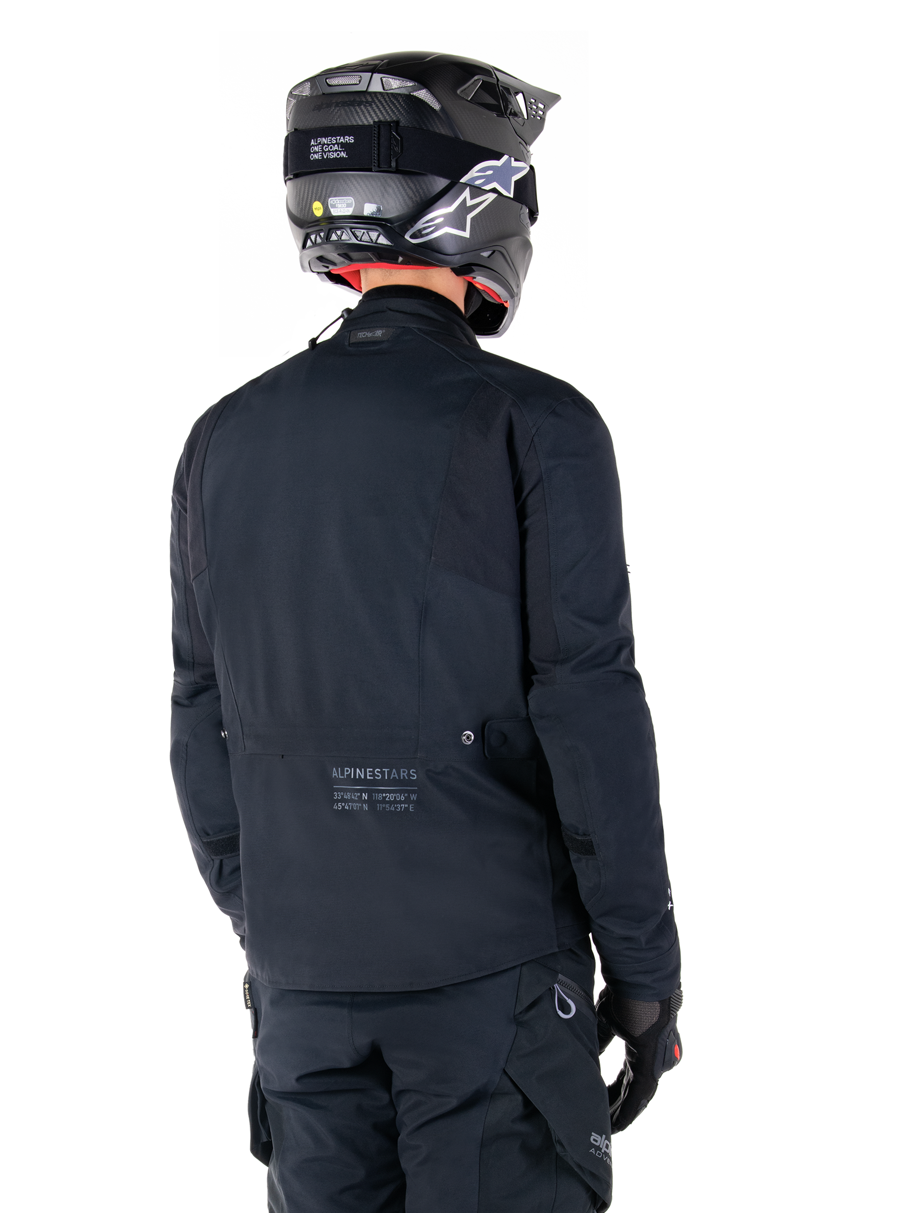 Alpinestars St-7 Prime GORE-TEX Jacke, Schwarz, Rückansicht eines Fahrers, der die Jacke mit passendem Helm und Hose trägt, mit Alpinestars-Branding und geografischen Koordinaten am unteren Rücken.