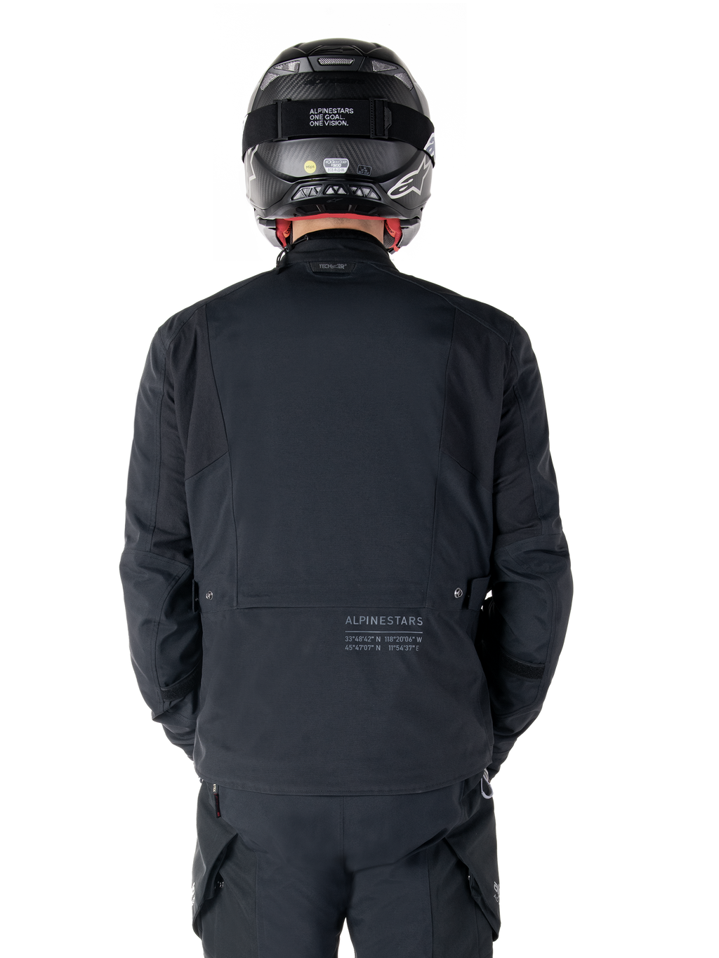 Alpinestars St-7 Prime GORE-TEX Jacke, Schwarz, Rückansicht eines Fahrers, mit dezentem Logo und geografischen Koordinaten auf dem unteren Rücken, getragen mit passendem schwarzem Helm und Hose, elegante und technische Motorradbekleidung