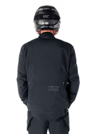Alpinestars St-7 Prime GORE-TEX Jacke, Schwarz, Rückansicht eines Fahrers, mit dezentem Logo und geografischen Koordinaten auf dem unteren Rücken, getragen mit passendem schwarzem Helm und Hose, elegante und technische Motorradbekleidung