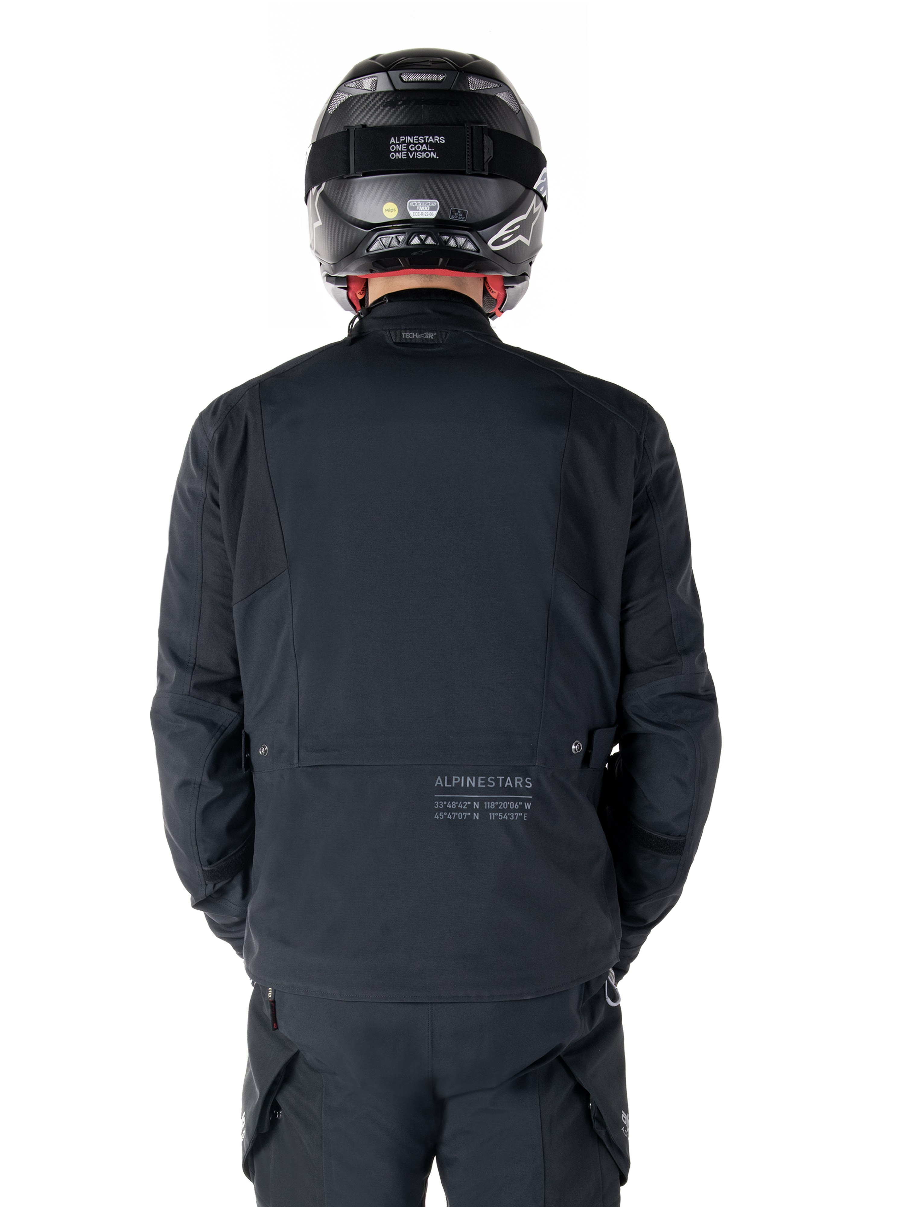 Alpinestars St-7 Prime GORE-TEX Jacke, Schwarz, Rückansicht eines Fahrers, mit dezentem Logo und geografischen Koordinaten auf dem unteren Rücken, getragen mit passendem schwarzem Helm und Hose, elegante und technische Motorradbekleidung
