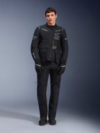 Alpinestars ST-7 2L GORE-TEX jas, adventure touring motorjas, zwart en donkergrijs, gedragen met bijpassende zwarte motorbroek en handschoenen, voorzien van meerdere cargozakken, reflecterende accenten en Alpinestars Exploration Equipment-logo op de borst, functioneel ontwerp voor rijden op de weg en off-road