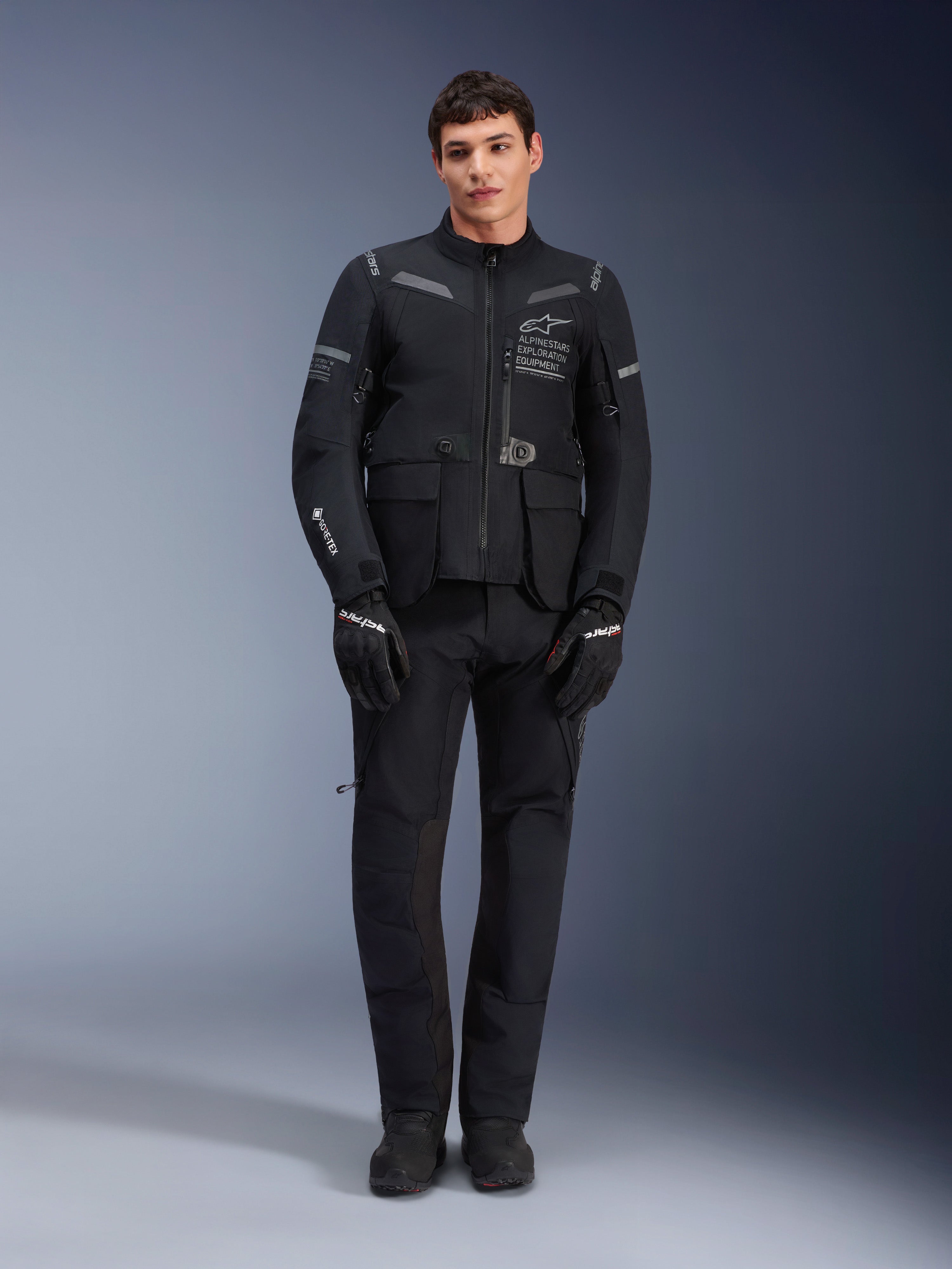 Alpinestars ST-7 2L GORE-TEX jas, adventure touring motorjas, zwart en donkergrijs, gedragen met bijpassende zwarte motorbroek en handschoenen, voorzien van meerdere cargozakken, reflecterende accenten en Alpinestars Exploration Equipment-logo op de borst, functioneel ontwerp voor rijden op de weg en off-road