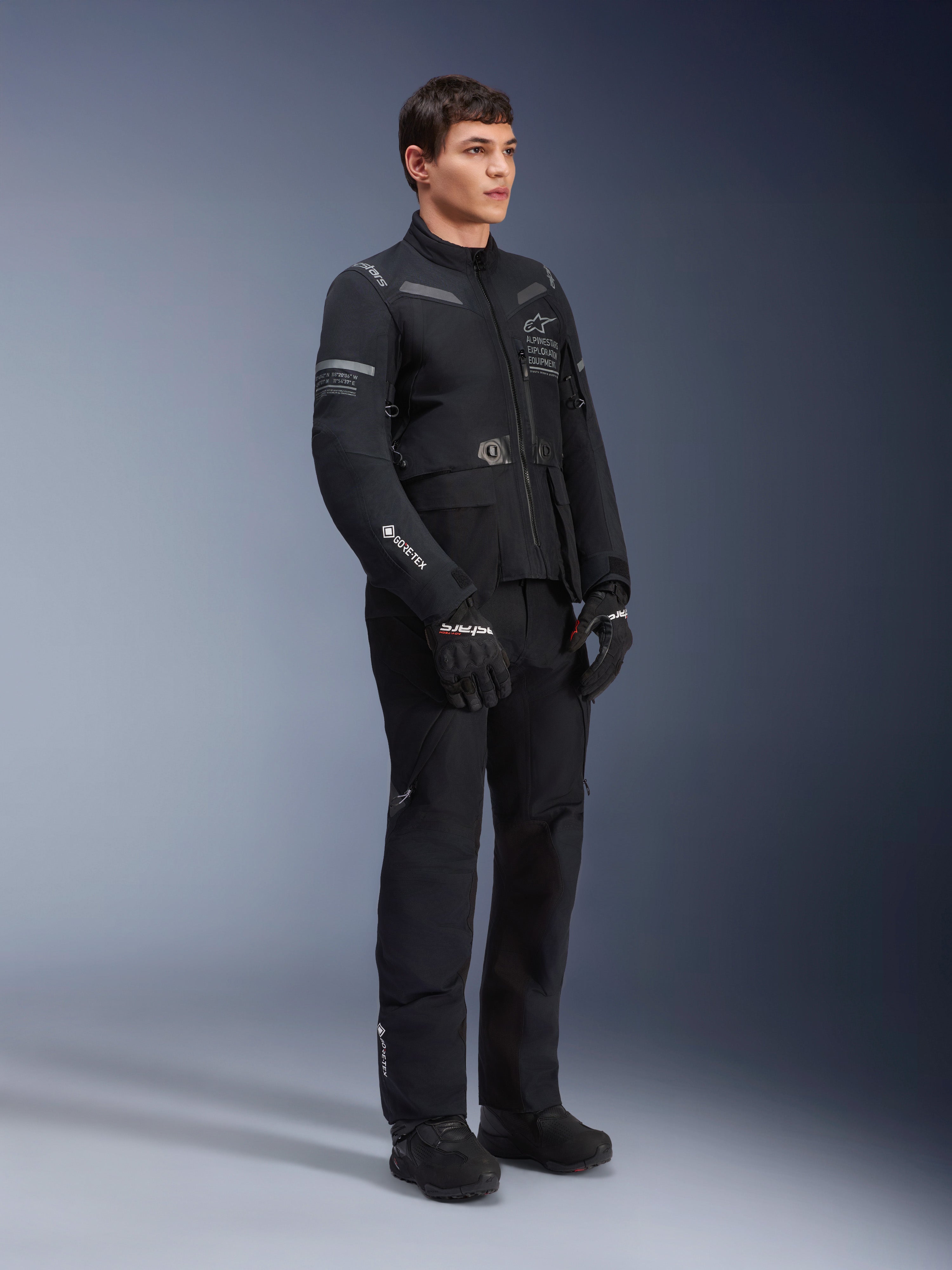 Mannelijk model draagt de Alpinestars ST-7 2L GORE-TEX jas in Black Dark Gray, adventure touring motorjas, bijpassende zwarte broek en handschoenen, driekwart aanzicht van het hele lichaam, voorzien van GORE-TEX logo en Alpinestars Exploration Equipment branding