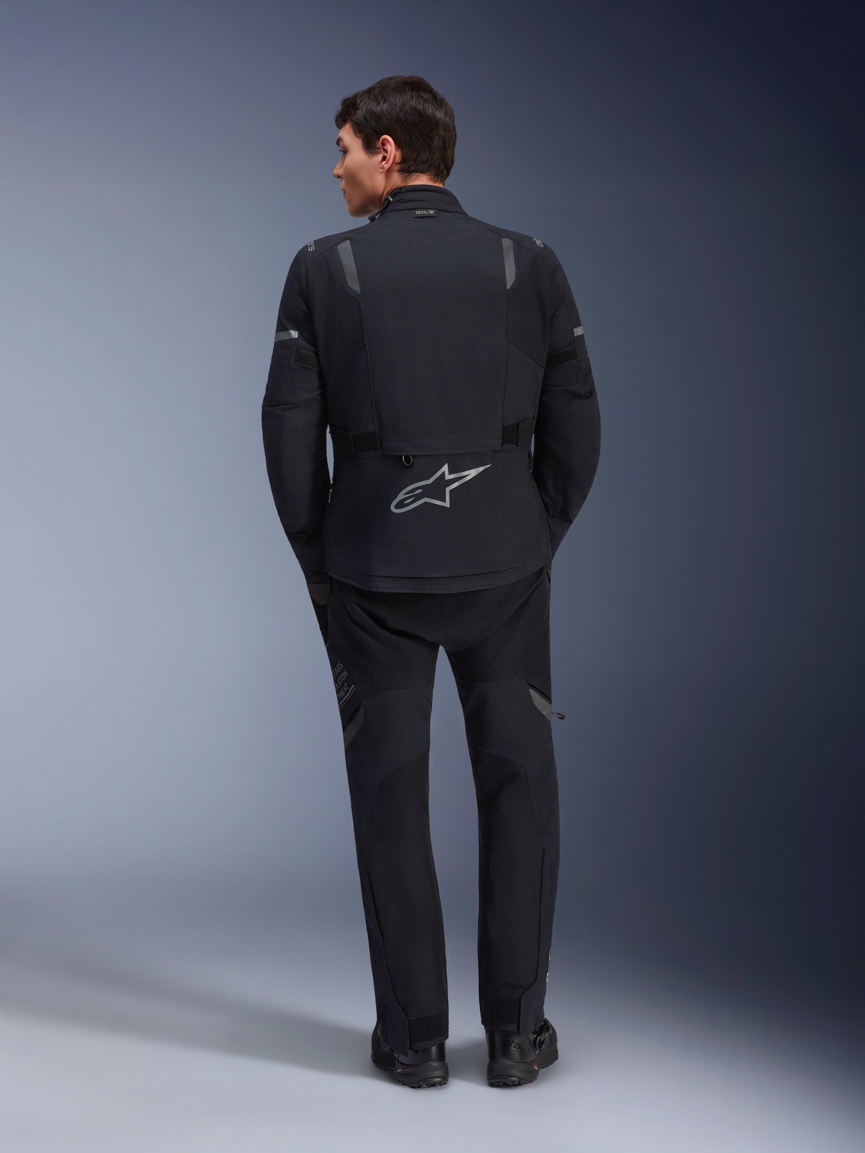 Alpinestars ST-7 2L GORE-TEX jas en bijbehorende ST-7 2L GORE-TEX broek, zwarte en donkergrijze adventure motoruitrusting, achteraanzicht met reflecterend Alpinestars-logo op de jas en technische textielconstructie