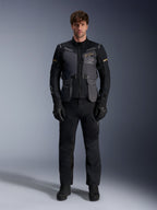 Mannelijk model draagt de Alpinestars ST-7 2L GORE-TEX Jas in Deep Gray Black Dark Khaki, voorzien van 'ALPINESTARS EXPLORATION EQUIPMENT' branding op de borst, gecombineerd met bijpassende zwarte motorbroek, handschoenen en laarzen, studio vooraanzicht.