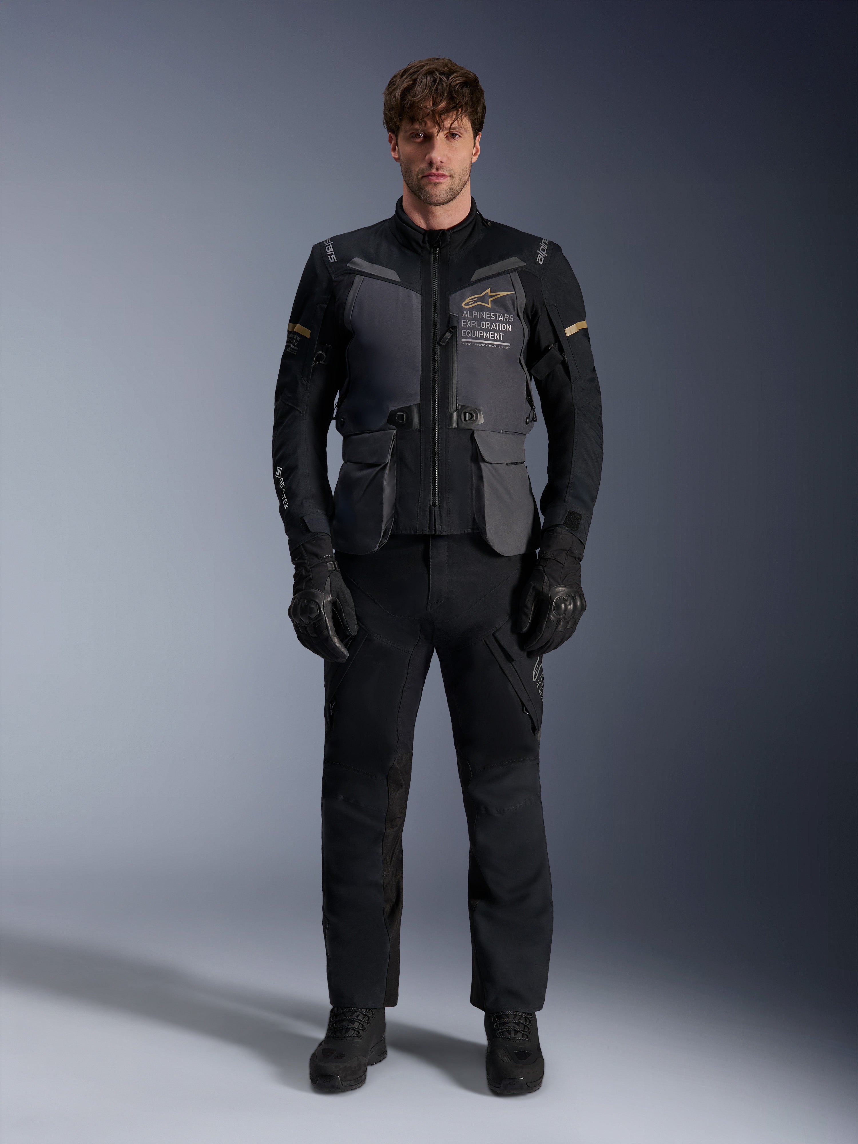 Mannelijk model draagt de Alpinestars ST-7 2L GORE-TEX Jas in Deep Gray Black Dark Khaki, voorzien van 'ALPINESTARS EXPLORATION EQUIPMENT' branding op de borst, gecombineerd met bijpassende zwarte motorbroek, handschoenen en laarzen, studio vooraanzicht.