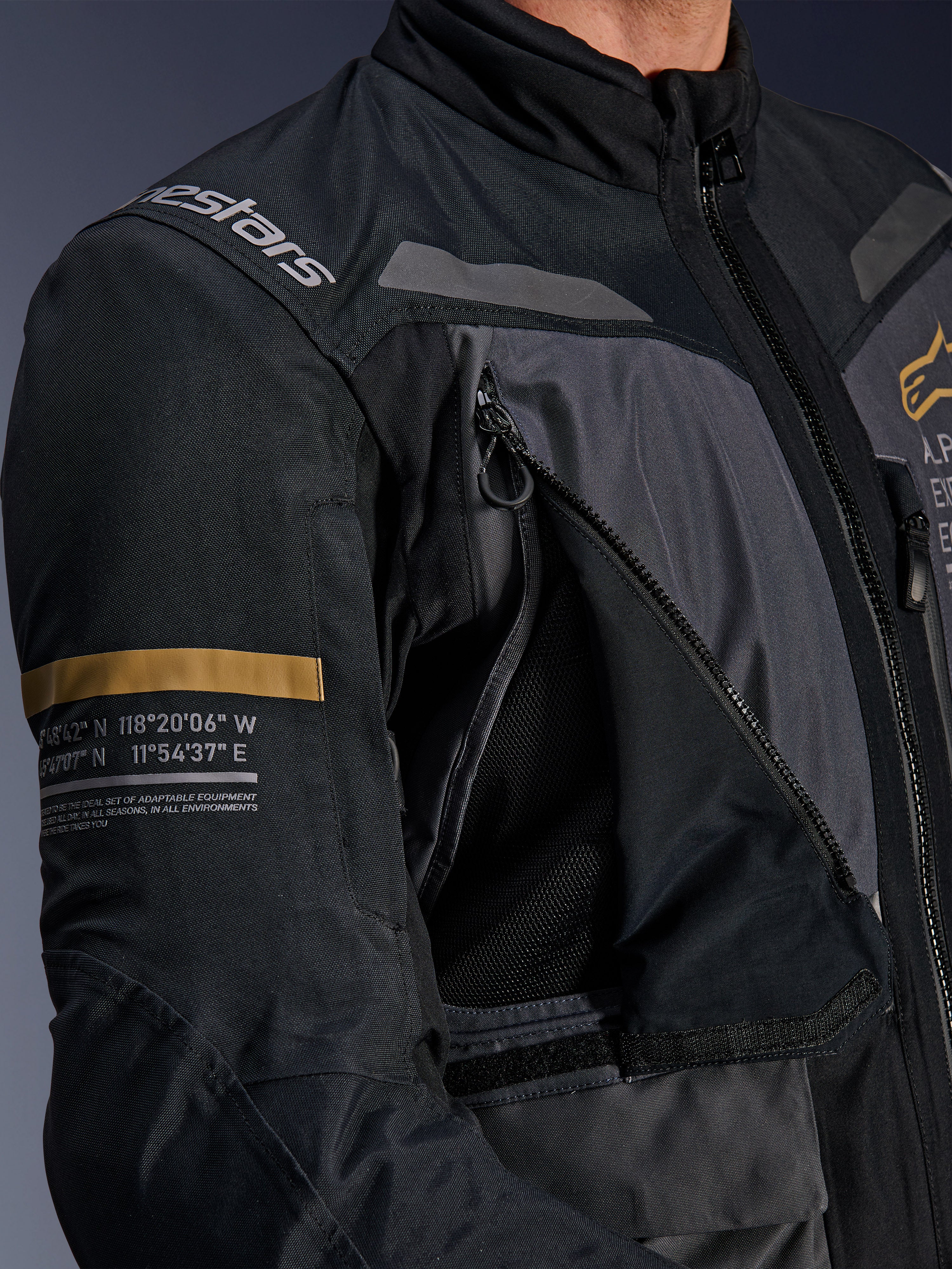 Alpinestars ST-7 2L GORE-TEX Jas, donkergrijs en zwart, close-up van geopend ventilatiepaneel op de borst met rits en mesh, Alpinestars-logo op de schouder en kaki streep op de mouw met geprinte geografische coördinaten, technische adventure-touring motoruitrusting