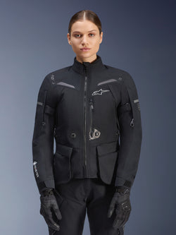 Frau in Alpinestars Stella ST-7 2L GORE-TEX Jacke, Schwarz mit dunkelgrauen Akzenten, Motorrad-Tourenjacke mit großen Cargo-Taschen und hohem Kragen, kombiniert mit schwarzen Handschuhen und Hosen, wasserdichte Adventure-Fahrbekleidung