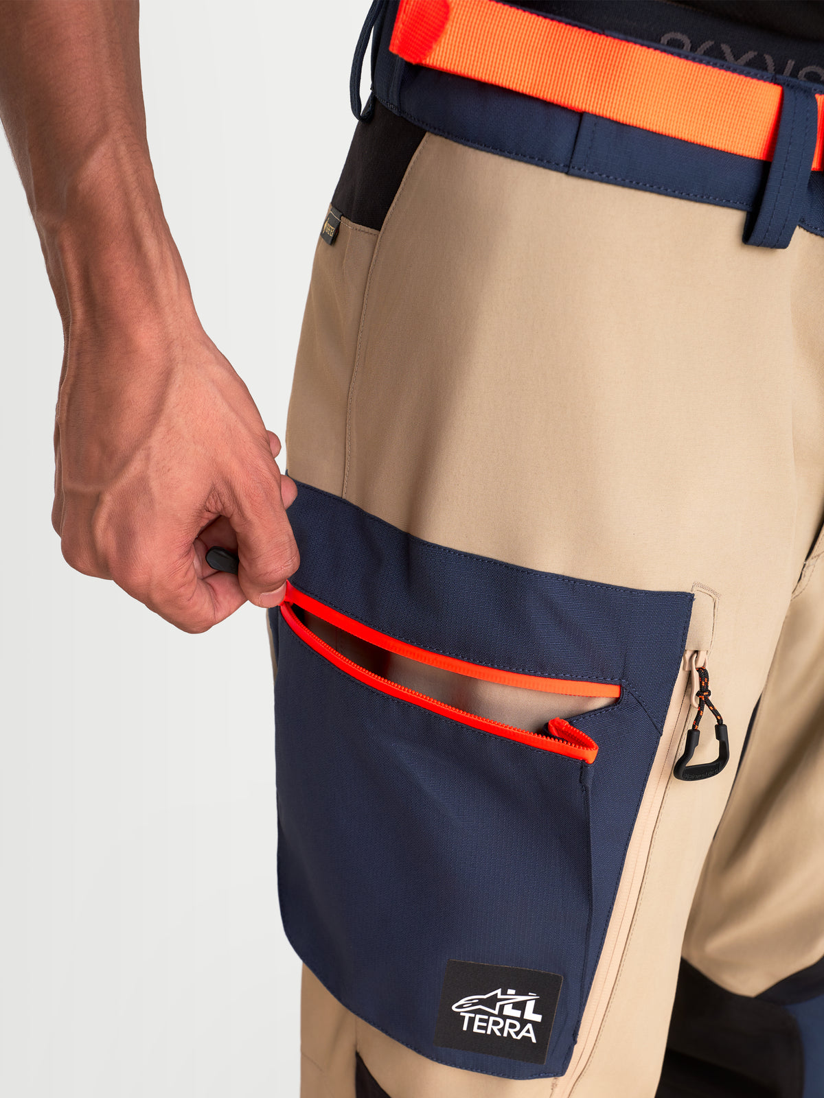 Alpinestars Arenis Gore-Tex Pantalons, pantalon de moto adventure, vue rapprochée montrant une main ouvrant la poche cargo intégrée avec des détails de zip orange vif, construction en textile étanche beige et bleu marine