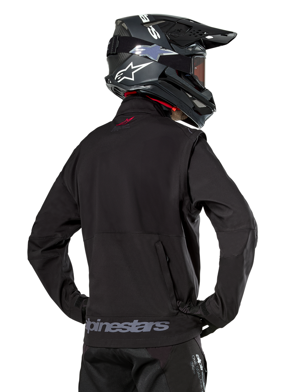 Veste Softshell Alpinestars Lite-Dura, noire, vue de dos portée par un pilote, avec marquage Alpinestars gris sur l'ourlet arrière et un petit logo rouge sur le col, associée à un casque de motocross noir, conçue pour l'enduro et la conduite tout-terrain.