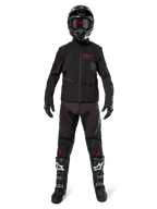 Pilote de motocross portant la veste softshell Alpinestars Lite-Dura noire avec des détails de logo rouges, associée à un pantalon tout-terrain noir assorti, un casque noir et des bottes blanches et noires, équipement complet de course enduro professionnel.