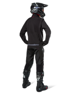 Pilote d'enduro vu de dos portant la veste softshell Alpinestars Lite-Dura en noir, associée à un pantalon technique et des bottes noirs, avec poche de rangement arrière zippée et logo Alpinestars sur l'ourlet inférieur, équipement tout-terrain complet pour le motocross.