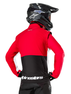 Veste Softshell Alpinestars Lite-Dura, Noir Rouge Vif Blanc, vue arrière d'un pilote de motocross, haut du corps et manches rouges avec empiècement noir en bas du dos, poche de rangement arrière zippée, logo Alpinestars blanc sur l'ourlet inférieur, portée avec un casque et un pantalon noirs