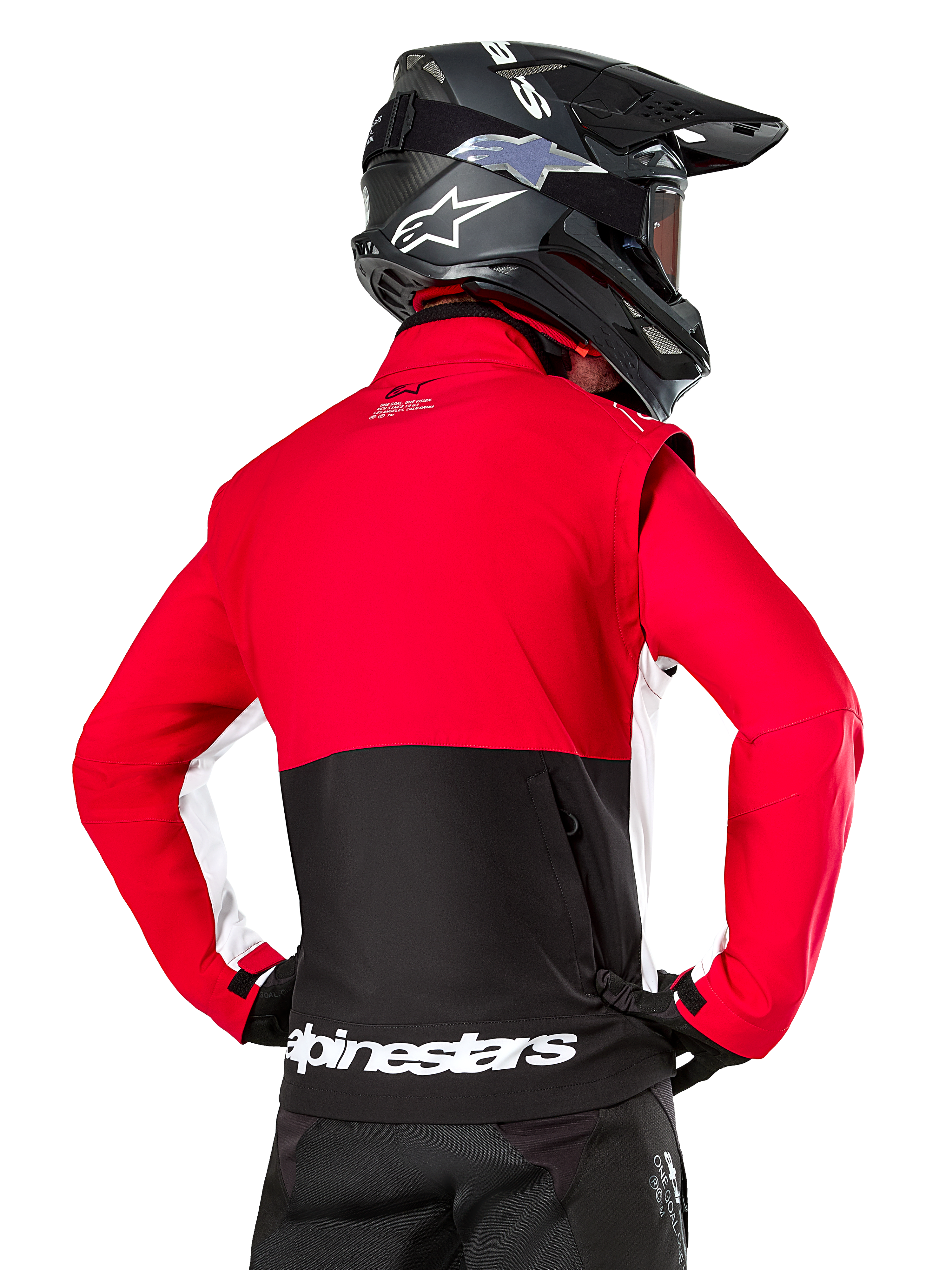 Veste Softshell Alpinestars Lite-Dura, Noir Rouge Vif Blanc, vue arrière d'un pilote de motocross, haut du corps et manches rouges avec empiècement noir en bas du dos, poche de rangement arrière zippée, logo Alpinestars blanc sur l'ourlet inférieur, portée avec un casque et un pantalon noirs