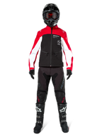 Pilote de motocross portant la veste Softshell Alpinestars Lite-Dura en noir, rouge vif et blanc, coordonnée avec un pantalon tout-terrain noir, des bottes noires et blanches et un casque noir avec lunettes, tenue complète pour la course d'enduro