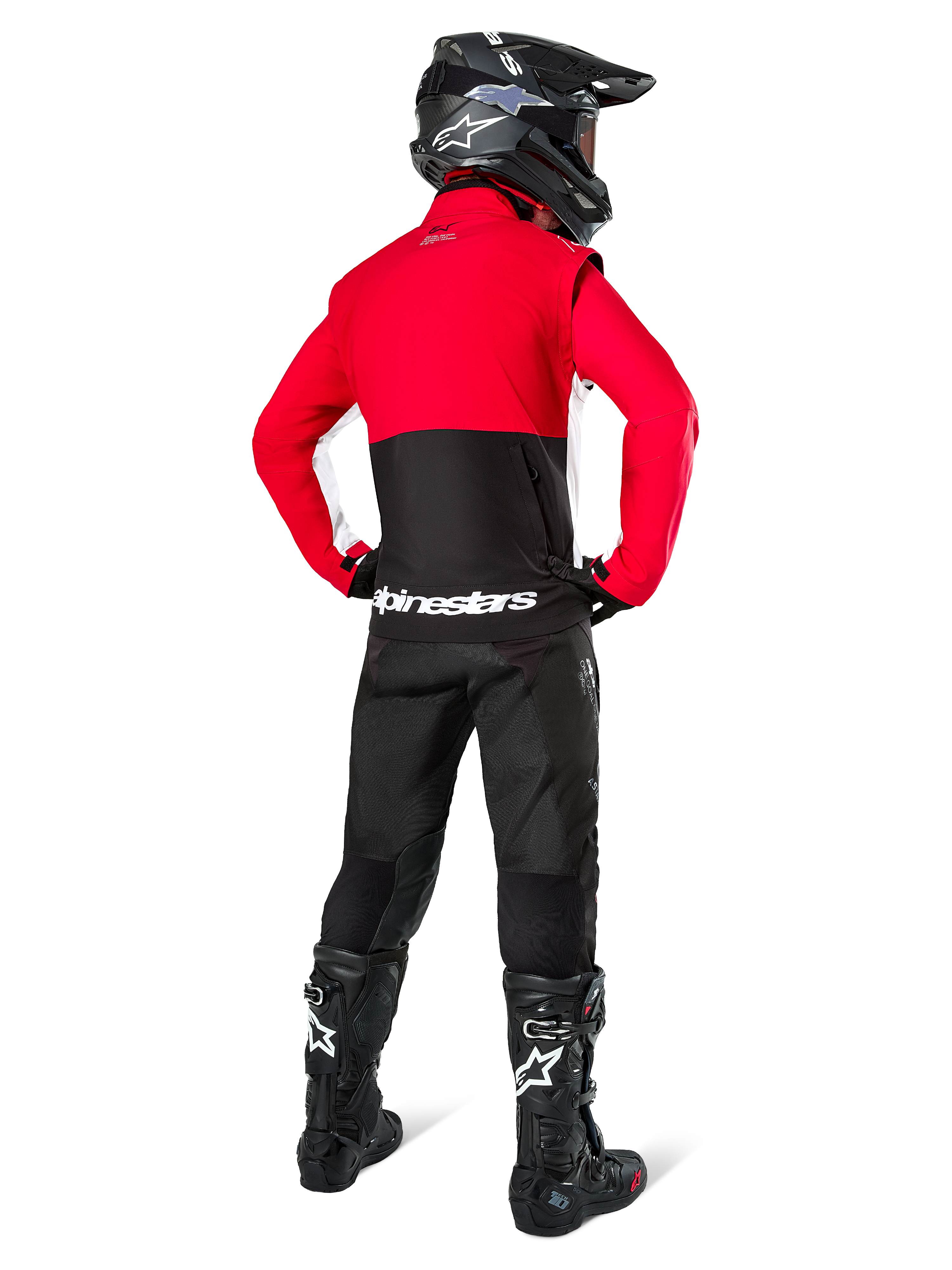 Pilote de motocross vu de dos portant la veste softshell Alpinestars Lite-Dura, noir, rouge vif et blanc, avec un empiècement supérieur rouge et une partie inférieure noire avec logo blanc, associée à un pantalon noir, un casque noir et des bottes noires, tenue d'enduro coordonnée pour la conduite tout-terrain