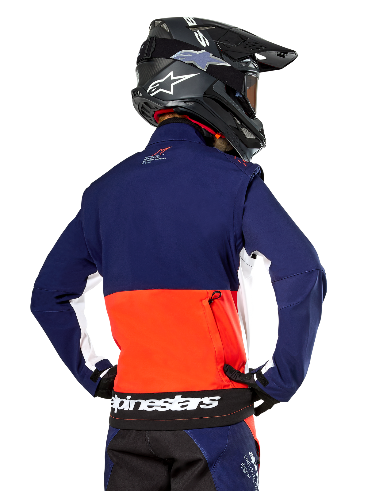 Veste Softshell Alpinestars Lite-Dura, bleu marine, orange vif et blanc, vue arrière portée par un pilote de motocross, avec un grand logo blanc sur l'ourlet arrière et une poche de rangement zippée, faisant partie d'une tenue de course tout-terrain complète.