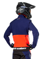 Veste Softshell Alpinestars Lite-Dura, bleu marine, orange vif et blanc, vue arrière portée par un pilote de motocross, avec un grand logo blanc sur l'ourlet arrière et une poche de rangement zippée, faisant partie d'une tenue de course tout-terrain complète.