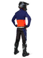 Pilote de motocross vu de dos portant la veste Softshell Alpinestars Lite-Dura en bleu marine, orange vif et blanc, montrant la grande poche de rangement arrière, avec un pantalon d'enduro assorti, un casque de motocross noir et des bottes tout-terrain noires