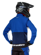 Alpinestars Lite-Dura Softshell Jacket in Navy, Bright Blue, and White, rear view showing the large zippered back pocket for sleeve storage, white side accents, and Alpinestars branding on the hem, worn with a black motocross helmet and matching gear.