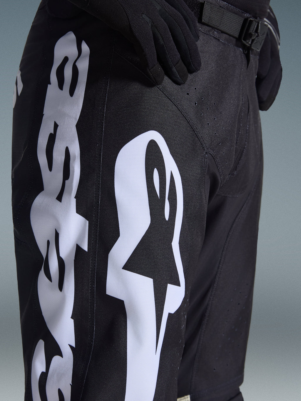 Alpinestars 2026 Racer Air Riway Pants, black motocross pants with bold white Alpinestars logo on the thigh, close-up showing technical fabric and adjustable waist strap, worn with black gloves for off-road riding