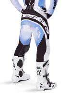 Alpinestars Pantalon Techstar Melt 2025, pantalon de motocross, noir, bleu et blanc, vue arrière avec logo Alpinestars imposant sur l'assise et logos étoiles sur les jambes, porté avec des bottes Tech 10 blanches et noires, équipement de course tout-terrain professionnel