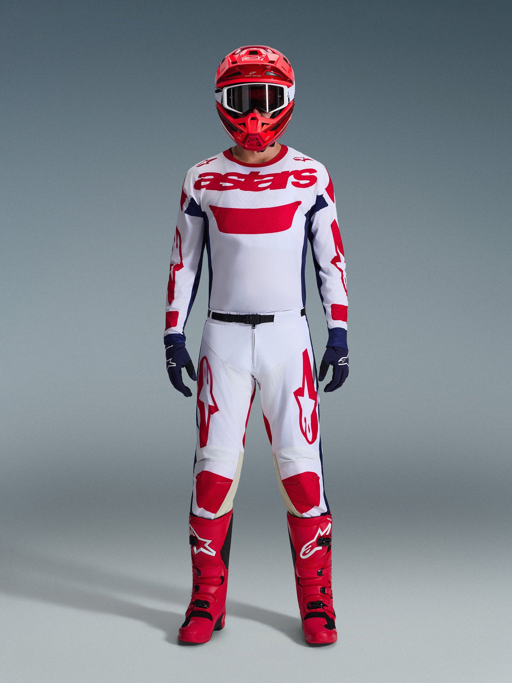Pilote de motocross portant l'ensemble d'équipement Alpinestars 2026 Racer Riway, maillot blanc avec marquage « astars » rouge et panneaux latéraux bleus, pantalon blanc avec graphismes rouges, casque de course et bottes rouges, gants bleu marine, tenue de motocross complète et coordonnée