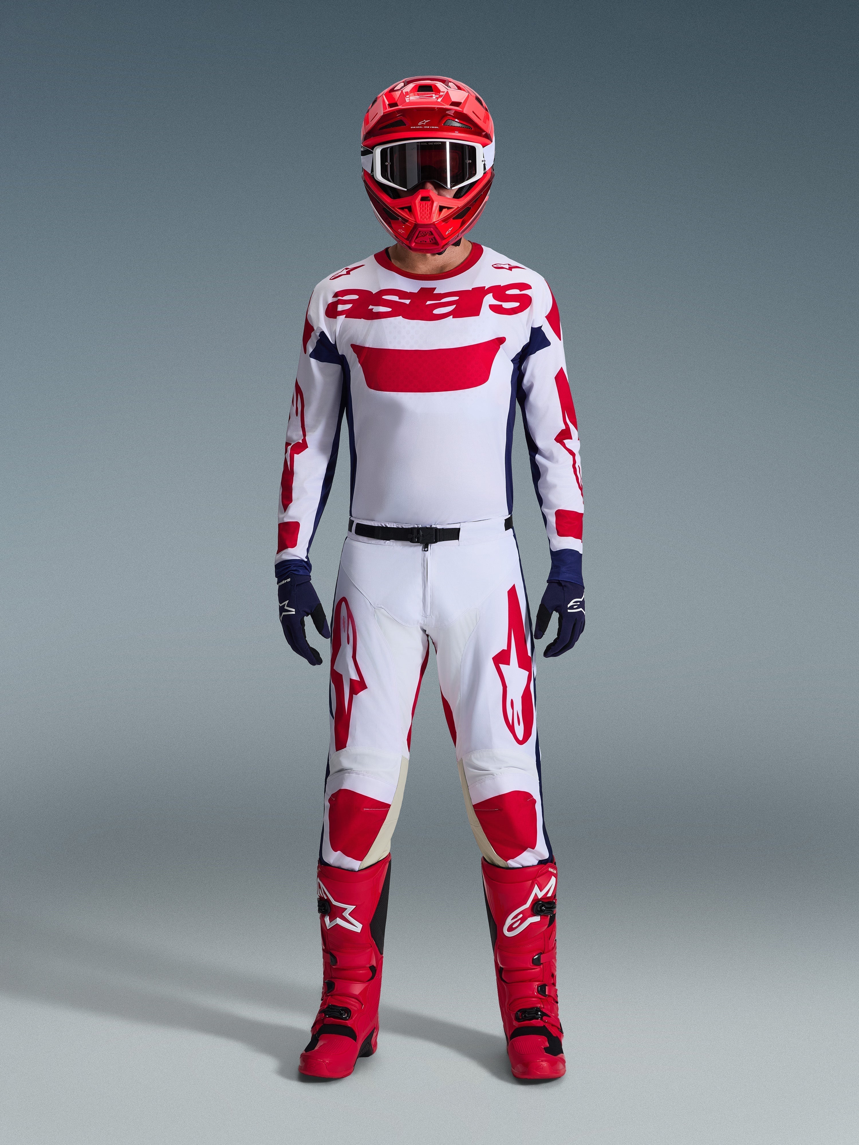 Pilote de motocross portant l'ensemble d'équipement Alpinestars 2026 Racer Riway, maillot blanc avec marquage « astars » rouge et panneaux latéraux bleus, pantalon blanc avec graphismes rouges, casque de course et bottes rouges, gants bleu marine, tenue de motocross complète et coordonnée