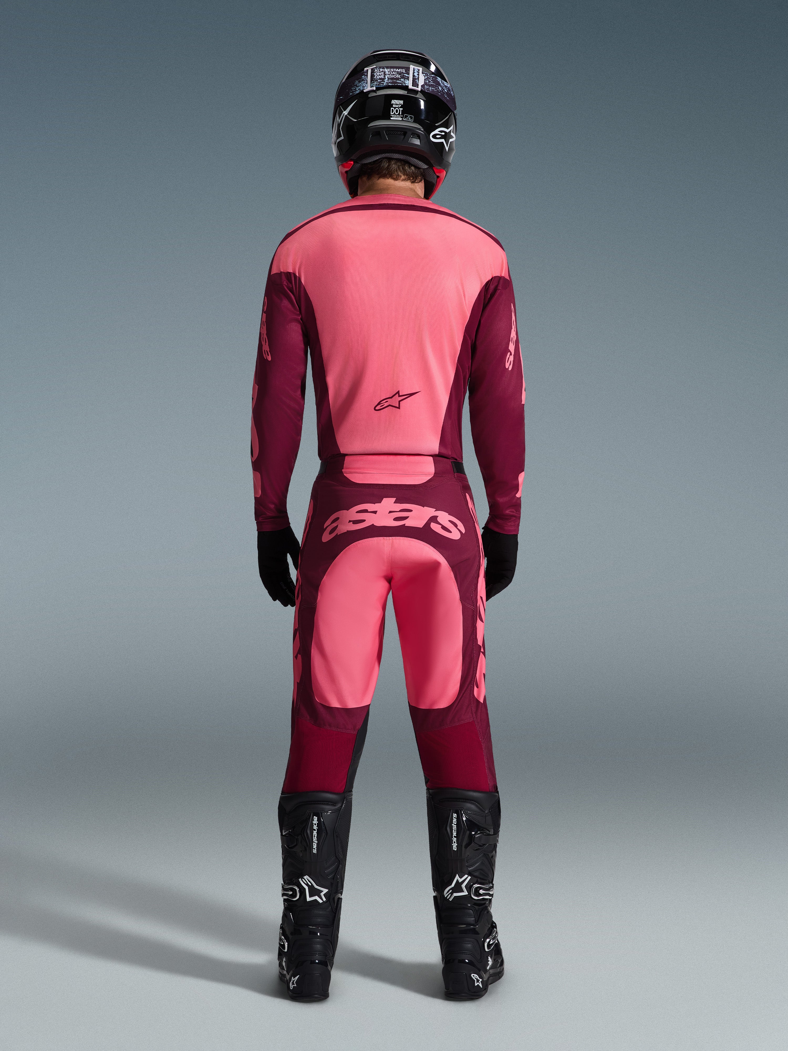 Rückansicht eines Motocross-Fahrers im 2026 Racer Riway Gear Set, pink- und bordeauxrotes Trikot, passende bordeauxrote Hose mit hellpinkem 'astars'-Logo auf dem Gesäß, schwarzer Helm, schwarze Stiefel und schwarze Handschuhe, abgestimmtes Outfit für Offroad-Performance