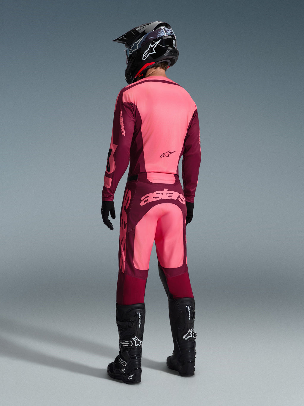 Motocross-Fahrer im Alpinestars 2026 Racer Riway Gear Set in der Rückansicht, Farbkombination in Kastanienbraun und Hellrosa, Trikot und Hose mit markantem Branding, komplett mit schwarzem Helm, Handschuhen und Stiefeln, abgestimmtes Motocross-Racing-Outfit