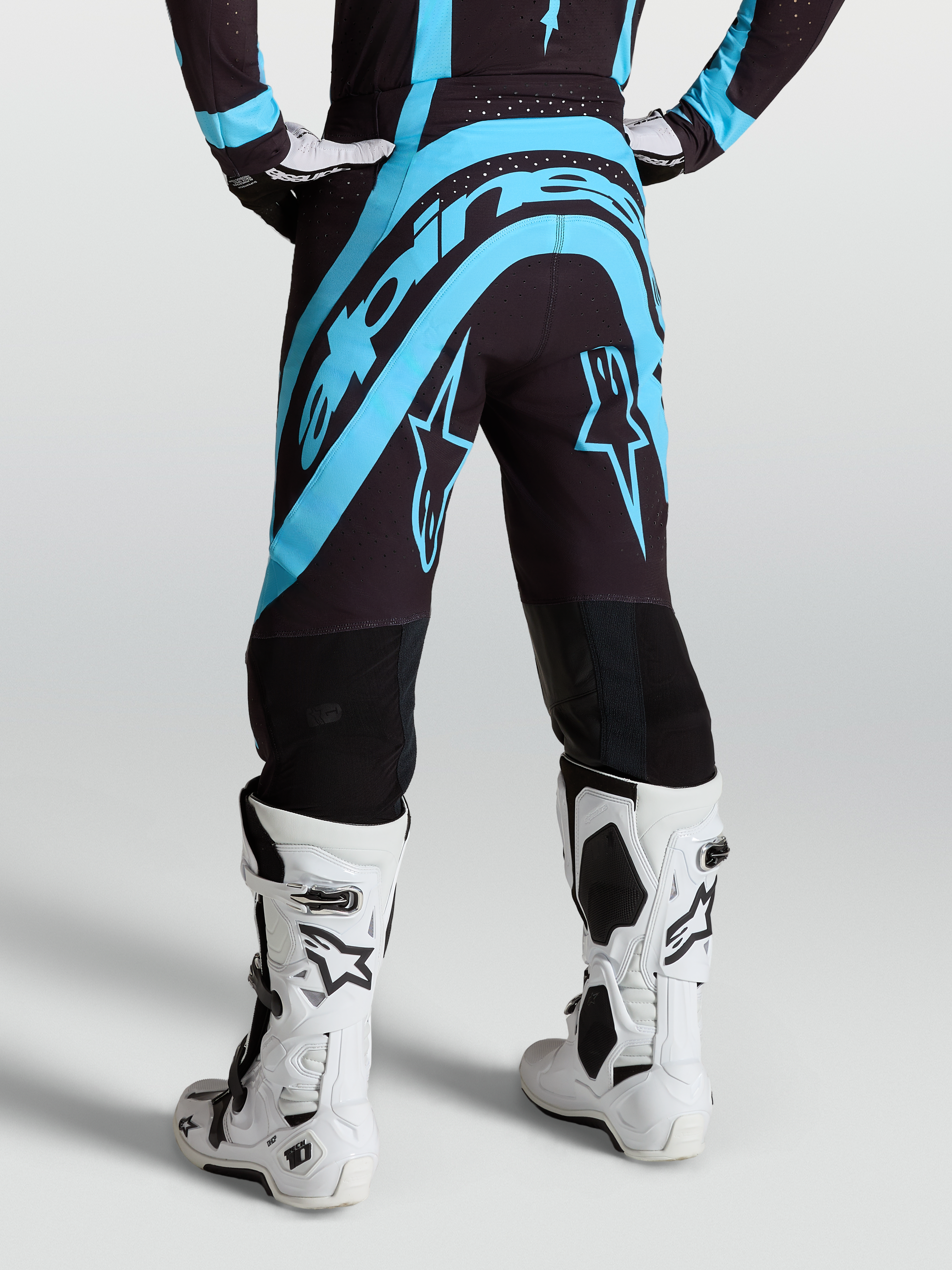 Alpinestars Supertech Vesta broek, professionele motocrossbroek in zwart en turquoise, achteraanzicht op model met witte motorlaarzen.