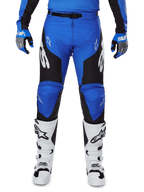 Alpinestars 2025 Racer Veil Pants, motorcrossbroek, blauw en zwart, met witte Alpinestars-logo's op de benen, zwarte zijpanelen, gedragen met witte motorcrosslaarzen en blauw/grijze handschoenen, hoogwaardige off-road rijuitrusting