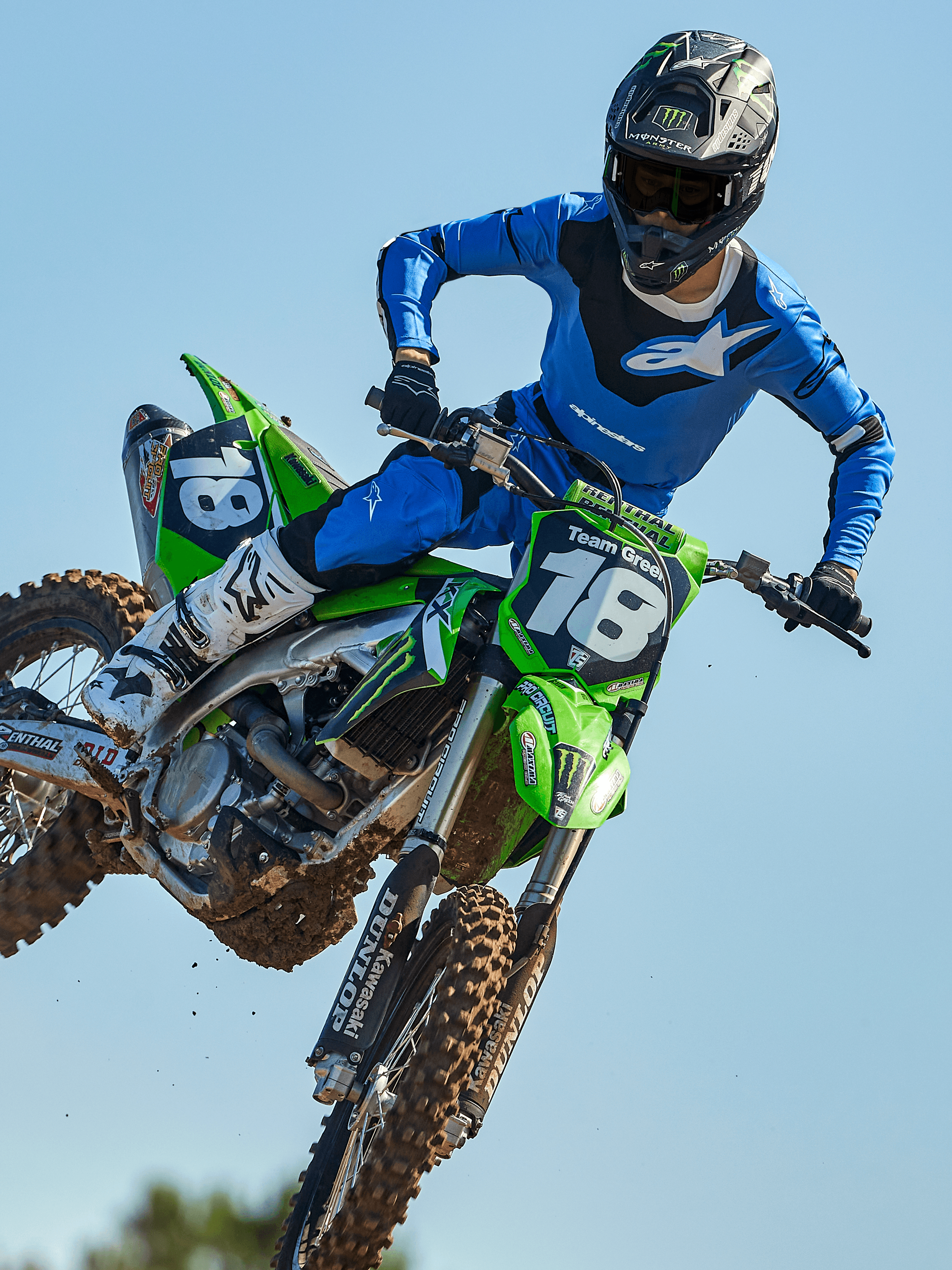 Motocrossrijder halverwege een sprong op een groene motorfiets, draagt Alpinestars 2025 Racer Veil Pants in Blauw en Zwart, bijpassende blauwe jersey met wit logo op de borst, witte en zwarte laarzen en zwarte helm, hoogwaardige off-road uitrusting voor racing.