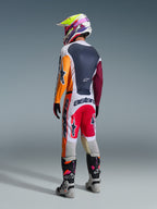Motocross rider viewed from behind wearing complete racing gear, Alpinestars 2026 Racer Portl Pants in Red Light Gray and Dark Gray, matching jersey with orange and maroon sleeves, multi-colored graphic helmet, gray and black off-road boots with pink highlights, professional motocross outfit