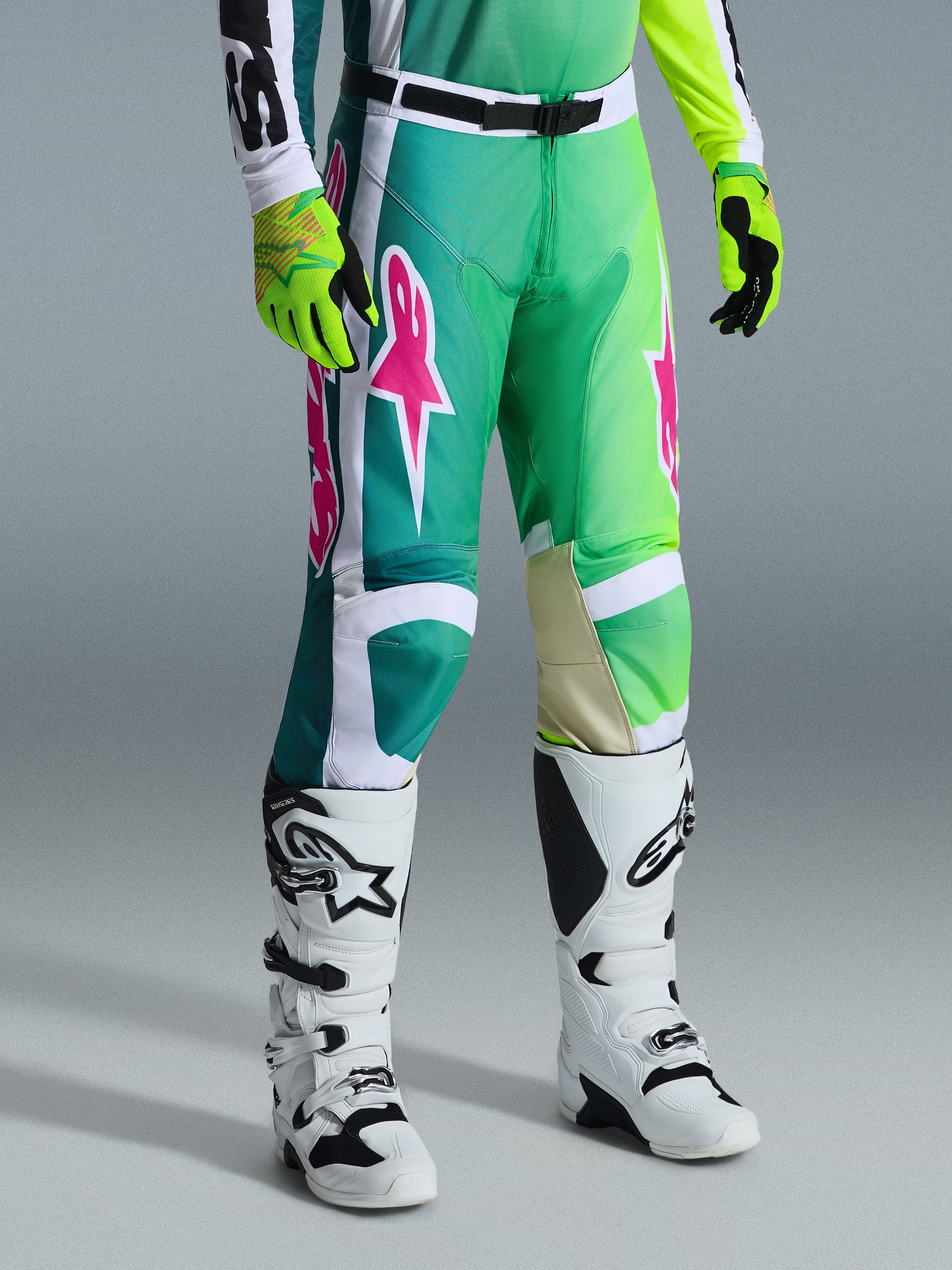 Alpinestars 2026 Racer Portl broek, motorcrossbroek, groen, wit en zwart met roze logo's, gedragen met witte laarzen en neongele handschoenen, voorzien van opvallende grafische patronen en een verstelbare zwarte taillegesp, prestatiegerichte uitrusting voor off-road rijden.