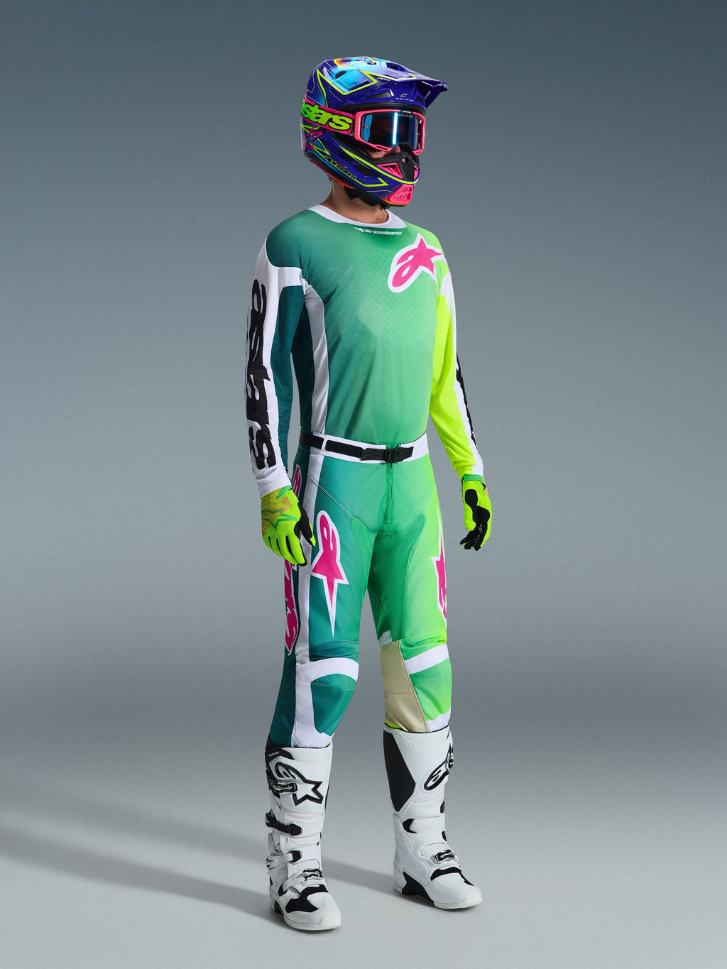 Motorcrossrijder met Alpinestars 2026 Racer Portl-broek en bijpassend shirt in groen, wit en zwart met roze logo-accenten, kleurrijke helm, witte en zwarte racelaarzen en neongroene handschoenen, complete kledingset voor off-road rijden