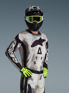 Motocrossrijder in de Alpinestars 2026 Racer Lahnd Jersey en kledingset, Sand Camo, lichtgrijs camouflagepatroon met zwarte logo's, zwarte helm met witte en neongele bril, fel neongele handschoenen, professionele off-road racekleding