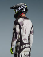Motorcrosser van achteren gezien in de Alpinestars 2026 Racer Lahnd Gear Set, grijs en wit Sand Camo jersey en broek met zwarte accenten, zwarte helm met wit Alpinestars-logo, neongele en zwarte handschoenen, gecoördineerde off-road performance outfit