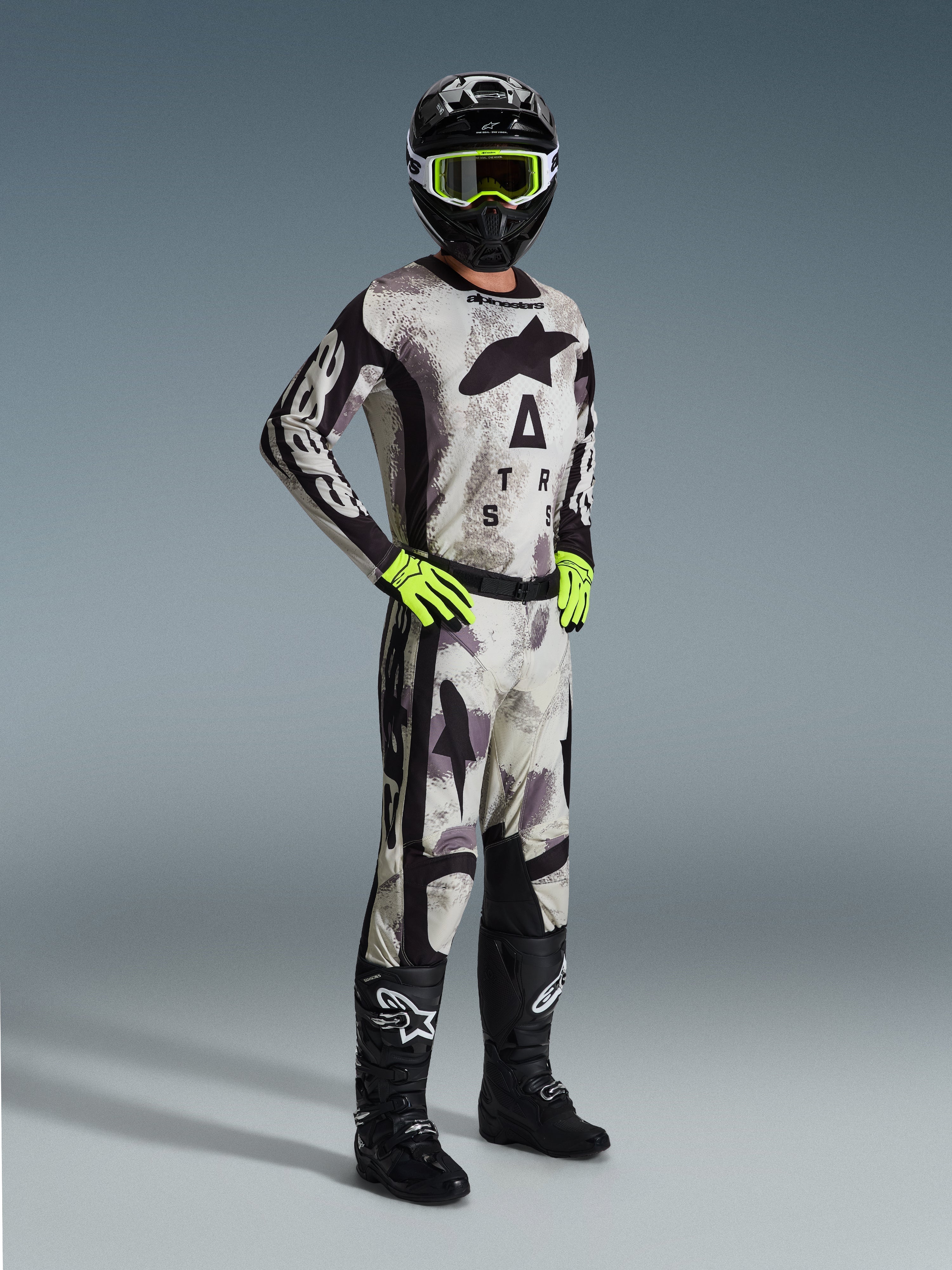 Alpinestars 2026 Racer Lahnd kledingset, motorcrossrijder in volledige race-uitrusting in Sand Camo en grijs, bijpassende jersey en broek met zwarte accenten en opvallende branding, neongele crossbril en handschoenen, zwarte helm en zwarte laarzen.