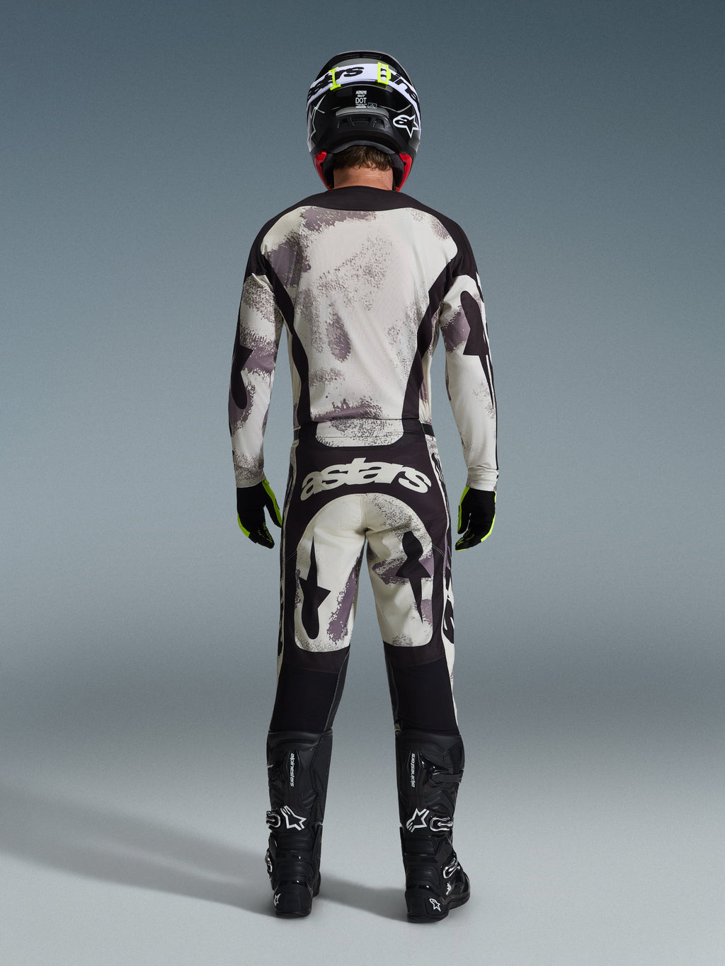 Pilote de motocross vu de dos portant le pantalon Alpinestars Racer Lahnd 2026 en Sand Camo, maillot à manches longues assorti avec motif camouflage gris, bottes de motocross noires, casque noir et marquage « astars » sur l'assise, conçu pour la course tout-terrain professionnelle.