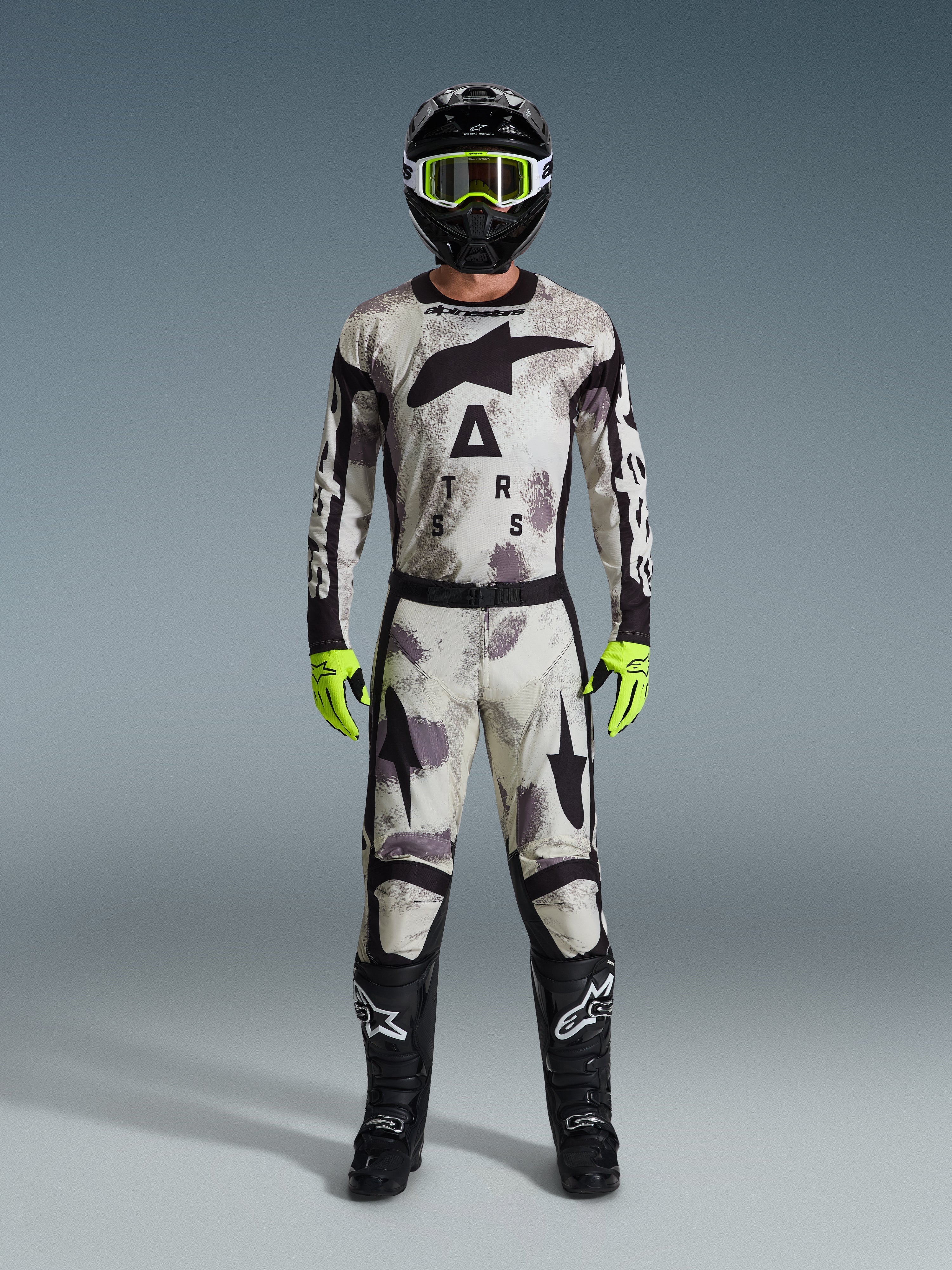 Pilote de motocross portant l'ensemble Alpinestars 2026 Racer Lahnd Gear Set, tenue à motif Sand Camo au design blanc, gris et noir, casque noir avec lunettes vert néon, gants vert néon éclatant et bottes noires avec logos blancs, tenue coordonnée complète pour la course tout-terrain