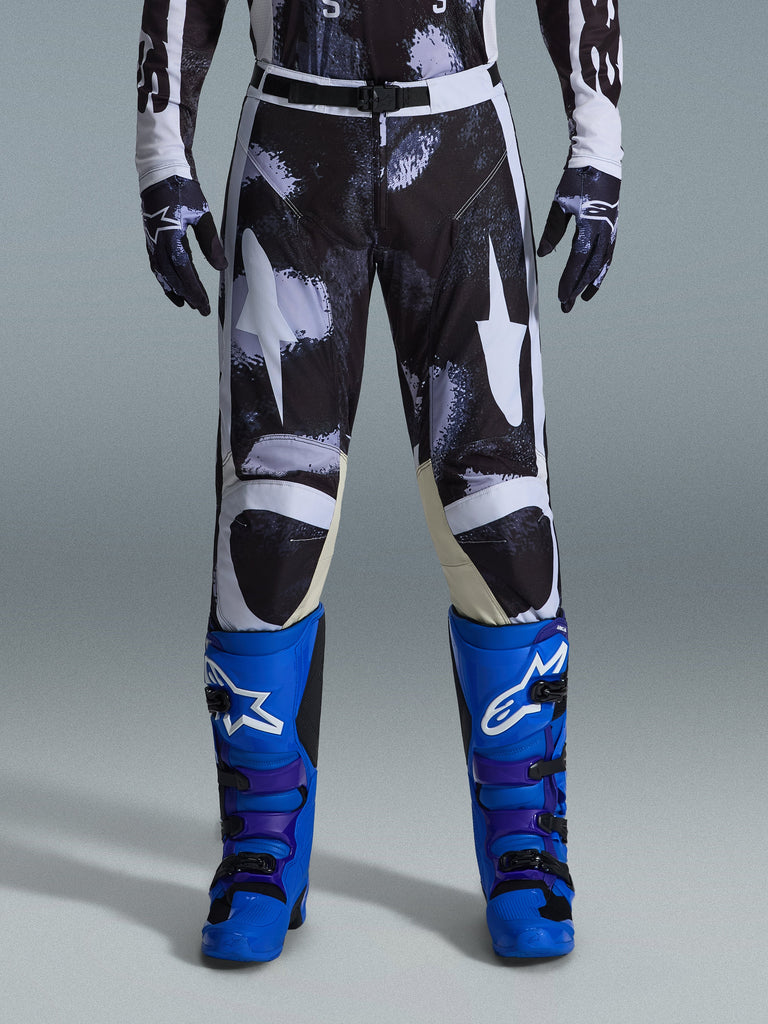 Alpinestars 2026 Racer Lahnd Pants, Motocross Pants, Iron Camo, worn with matching camouflage gloves and bright blue and purple boots, featuring stylized white star graphics on a dark camo pattern, technical magnetic buckle closure, professional off-road racing gear.