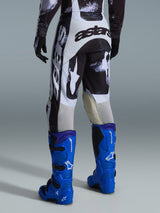 Pantalon Alpinestars Racer Lahnd 2026, pantalon de motocross, motif Iron Camo gris et noir, vue arrière montrant un large marquage astars blanc et noir, associé à des bottes de motocross bleu vif, équipement de course tout-terrain professionnel